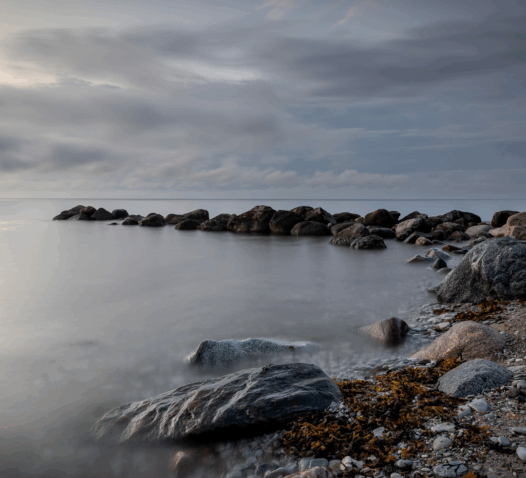 Kustlandskap med klippor och stilla vatten under en molnig himmel.